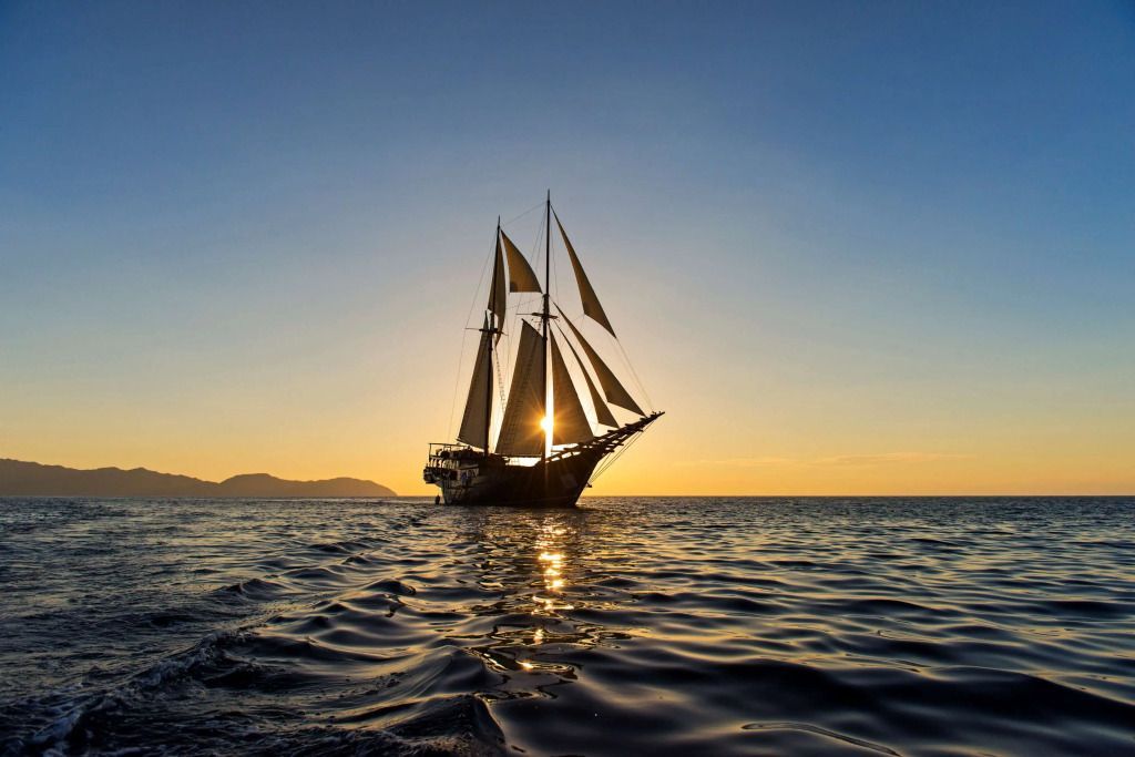 Segelschiff auf offenem Meer bei Sonnenuntergang mit Bergen im Hintergrund