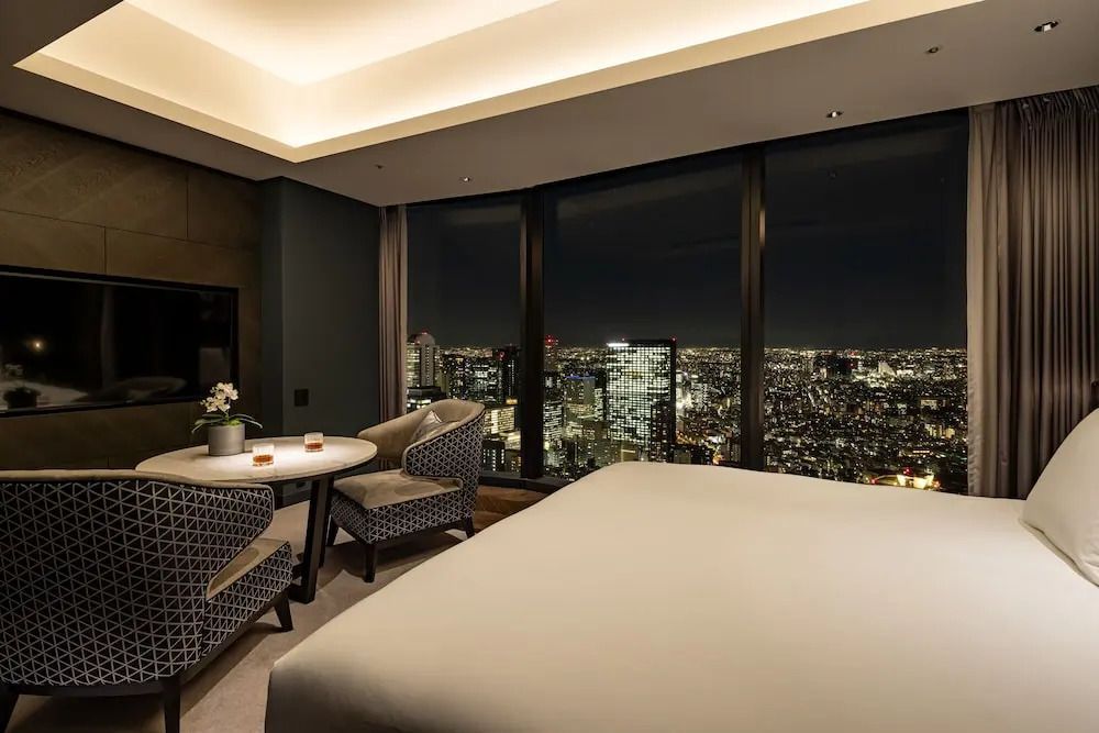 Nachtsicht aus einem Hotelzimmerfenster mit Stadtlichtern und Doppelbett im Vordergrund.