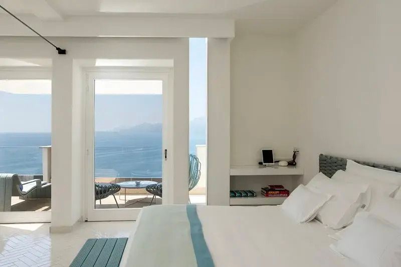 Zimmer mit großem Fenster und Aussicht auf das Meer und die Terrasse mit Sitzgelegenheiten.