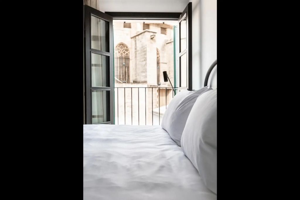 Bett mit weißer Bettwäsche und Blick durch Fenster auf historisches Gebäude.