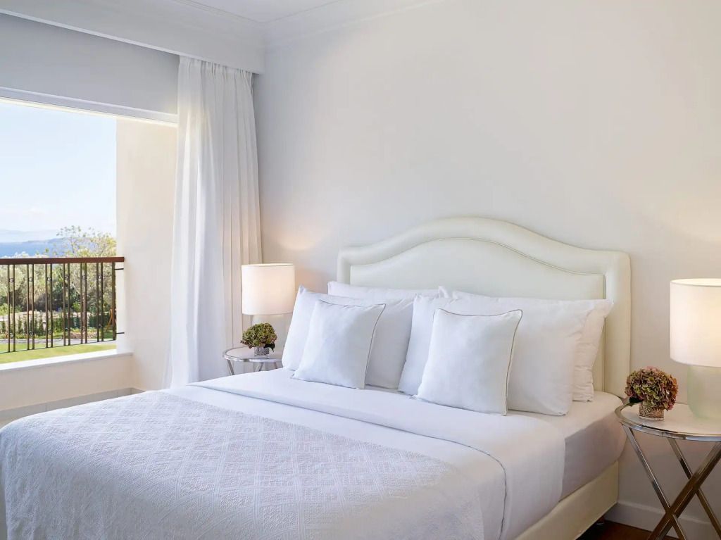 Zimmer mit einem weißen Doppelbett und Blick auf einen sonnigen Balkon