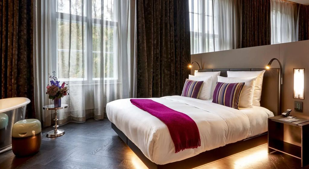 Helles Schlafzimmer mit großem Doppelbett, lila Akzent und Blick auf Vorhänge