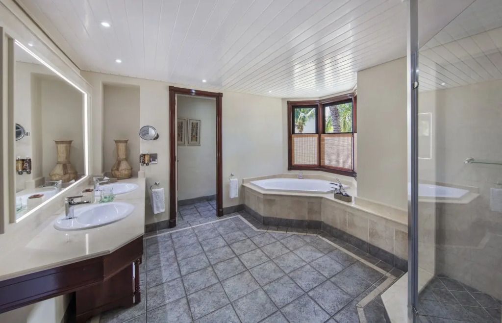 Badezimmer mit Eckbadewanne, zwei Waschbecken und eleganten Fensterjalousien.