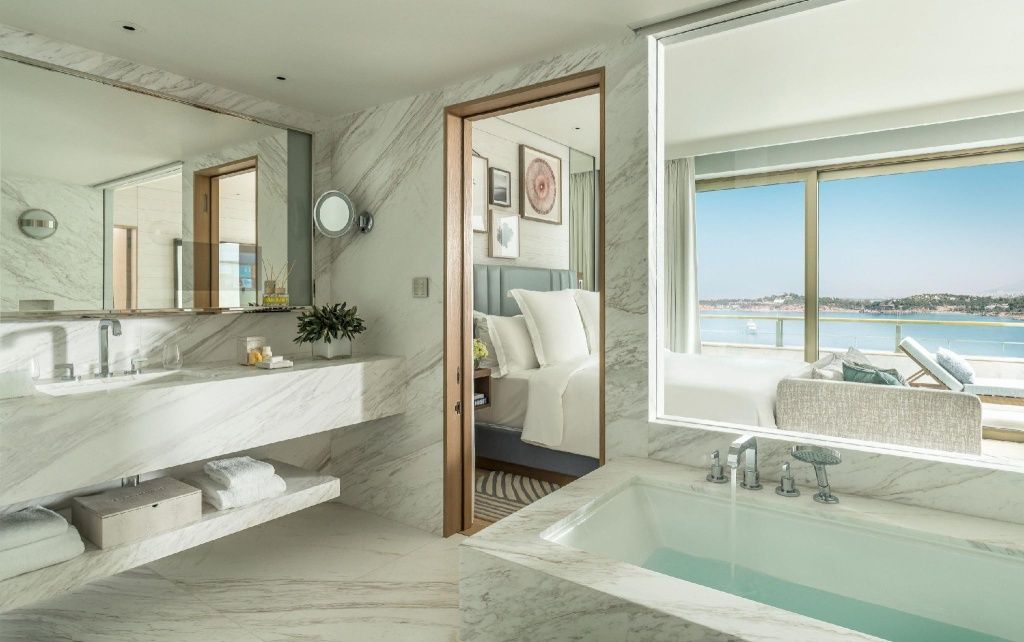 Luxuriöses Badezimmer aus weißem Marmor mit Badewanne und Blick auf das angrenzende Schlafzimmer.