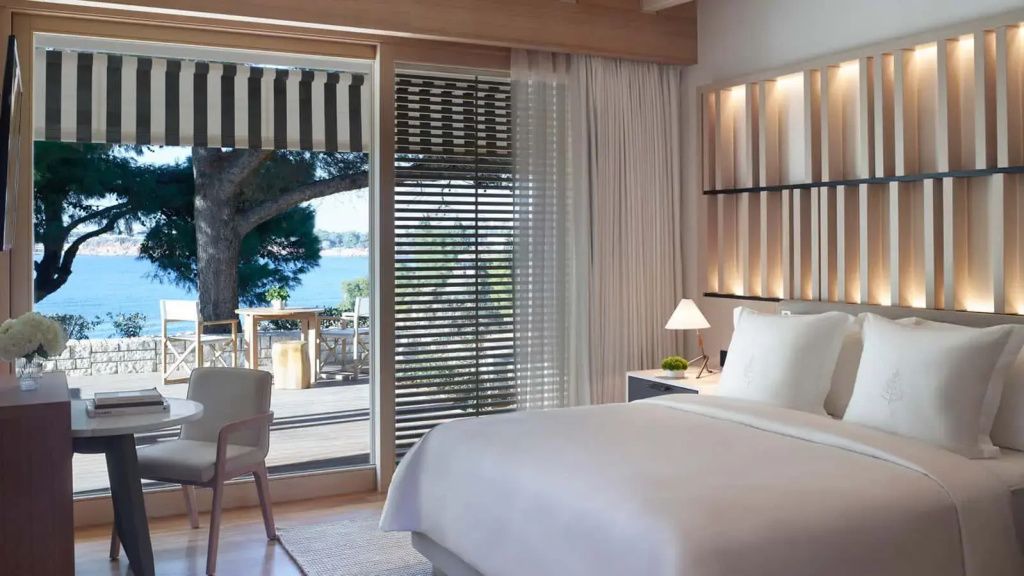 Doppelbett im Bungalow mit Blick durch große Glasfront auf die Terrasse und das Meer.