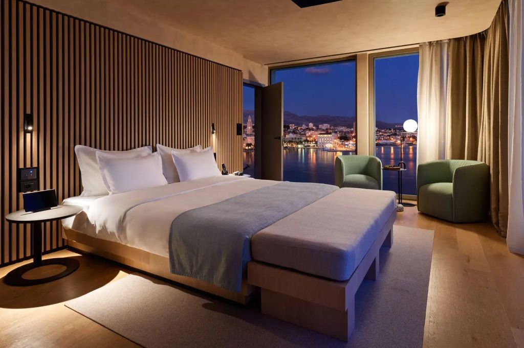 Abendliches Zimmer mit Doppelbett und beleuchteter Stadtansicht durch Fenster