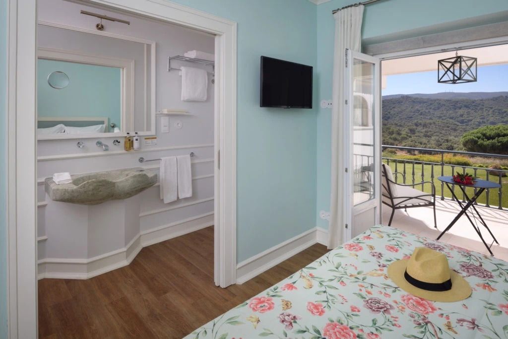 Zimmer mit pastellblauen Wänden, Blick ins Badezimmer und Zugang zum Balkon mit Bergsicht.