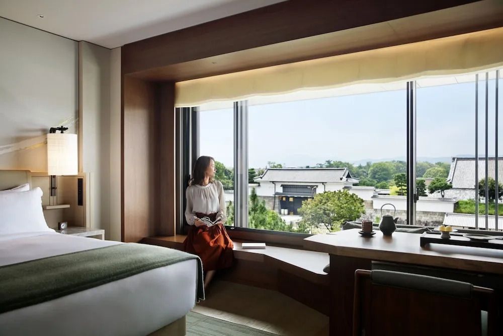 Frau sitzt am Fenster eines Hotelzimmers mit Blick auf grünen Außenbereich