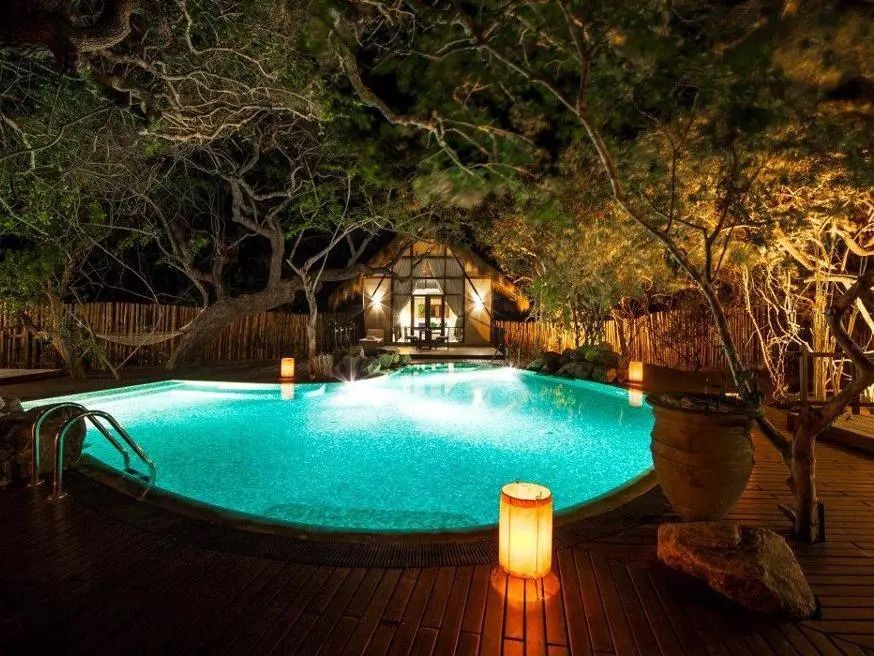 Beleuchteter Pool in tropischer Umgebung mit Holzdeck und Laternen bei Nacht.