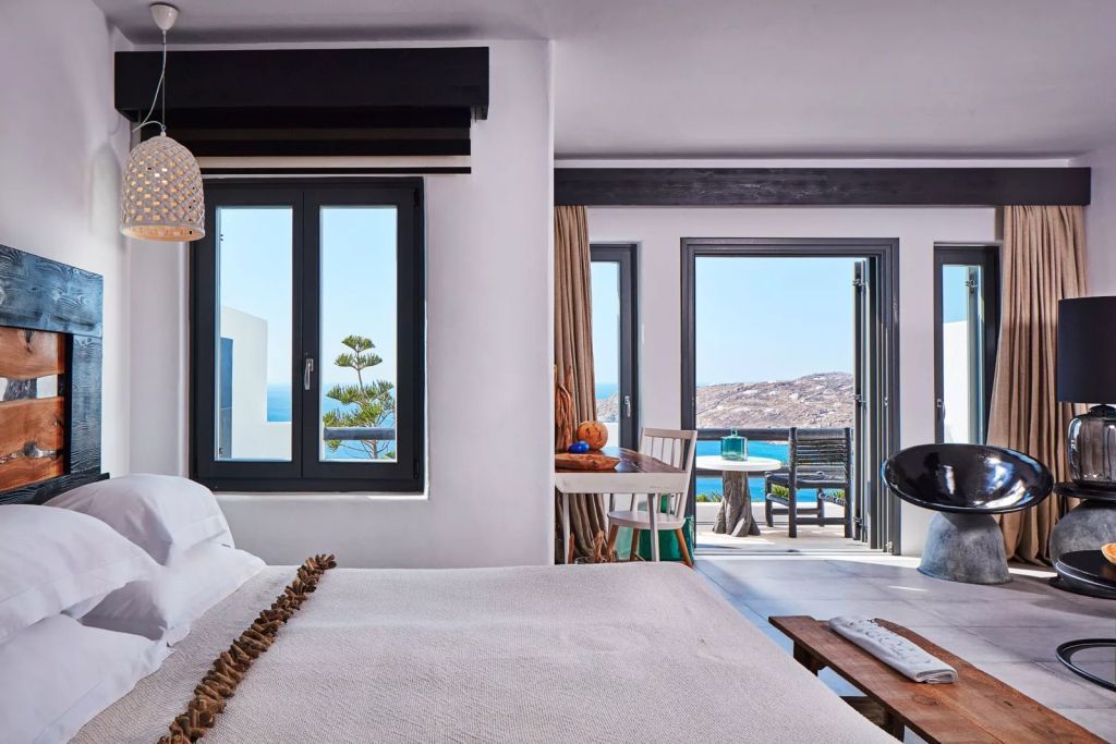Helles Zimmer mit Doppelbett, Meerblick von zwei großen Fenstern und kleinem Esstisch.