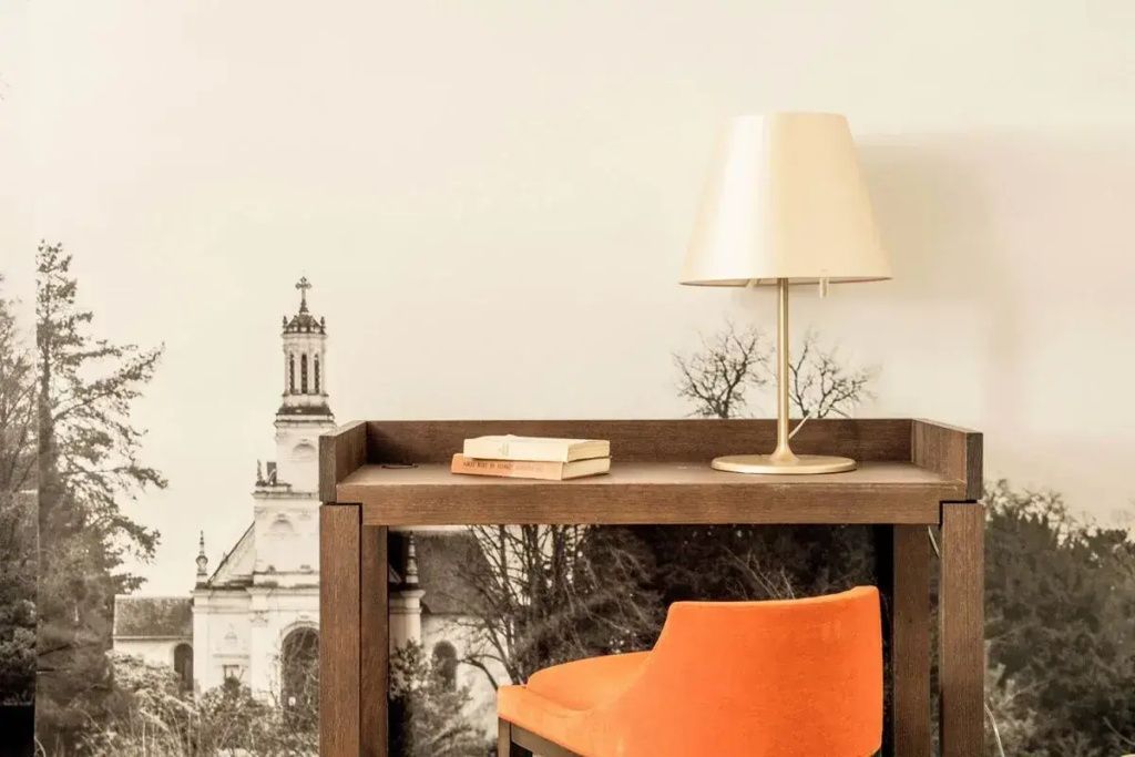 Schreibtisch mit orangem Stuhl und Lampe vor historischer Gebäudetapete.