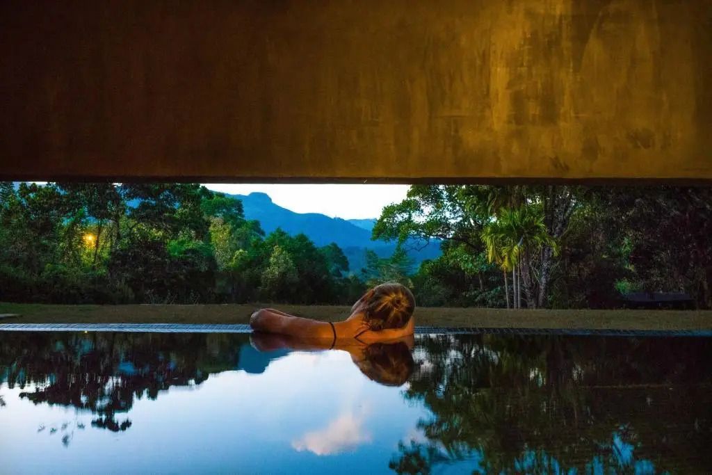 Person entspannt in einem Infinity-Pool mit Blick auf Wald und Berge bei Sonnenuntergang