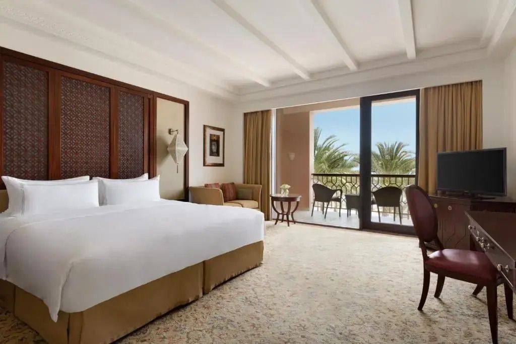 Deluxe Zimmer mit Balkon und Blick auf Palmen, großes Bett und gemütliche Sitzgelegenheit
