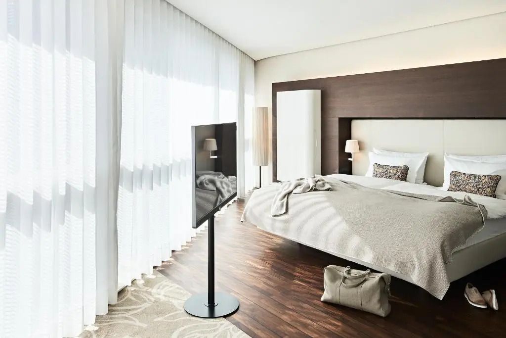 Modernes Hotelzimmer mit Doppelbett, Holzfußboden und großen Fenstern mit Vorhängen.