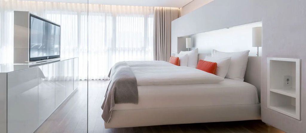 Modernes Doppelbett in heller Suite mit weißen und orangen Kissen.