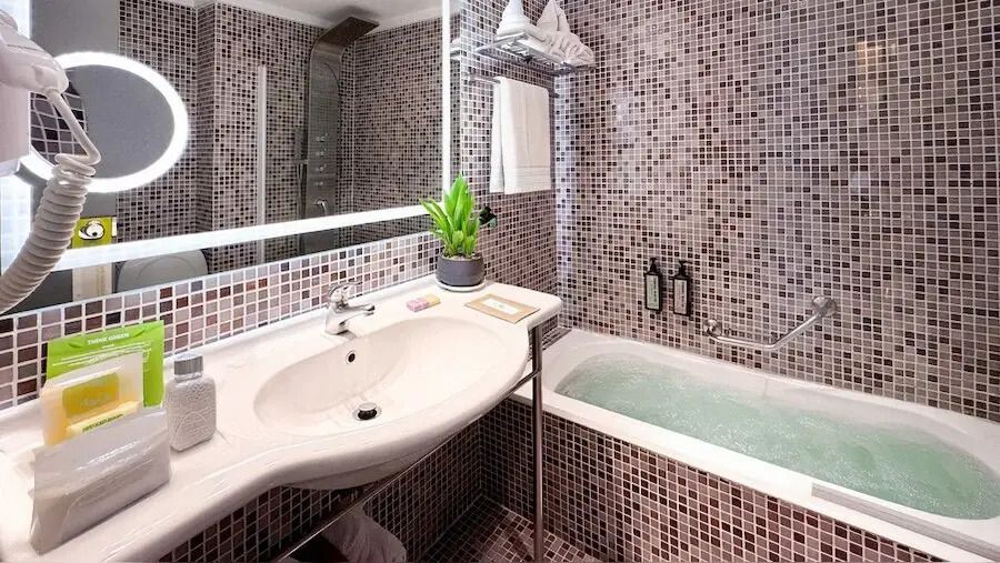 Modernes Badezimmer mit Mosaikfliesen, großem Spiegel und Badewanne mit frischem Grün.