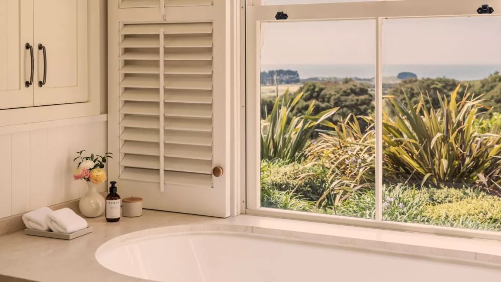 Badewanne mit Aussicht auf grüne Vegetation und Meer durch Fenster.