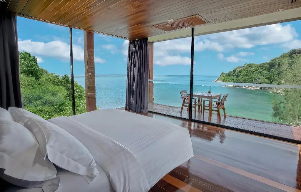 Modernes Schlafzimmer mit großen Fenstern und Meerblick sowie Balkon mit Tisch und Stühlen.