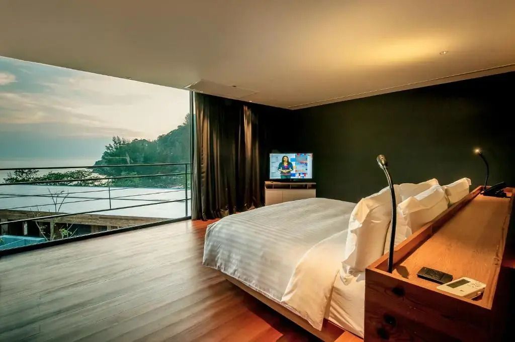Zimmer mit großem Bett, Balkon und Meerblick. Fernseher an der Wand, dunkle Vorhänge.