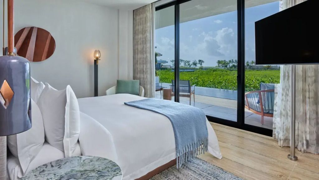 Helles Schlafzimmer mit Doppelbett und Blick auf tropische Vegetation durch große Fenster.