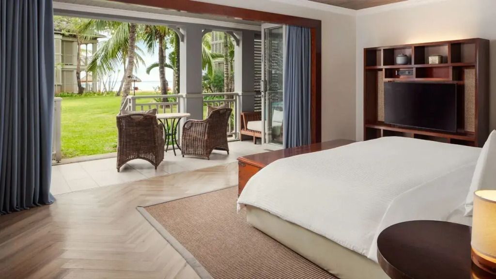 Junior-Suite mit Doppelbett und Blick auf eine Terrasse mit Gartenmöbeln und Palmen.