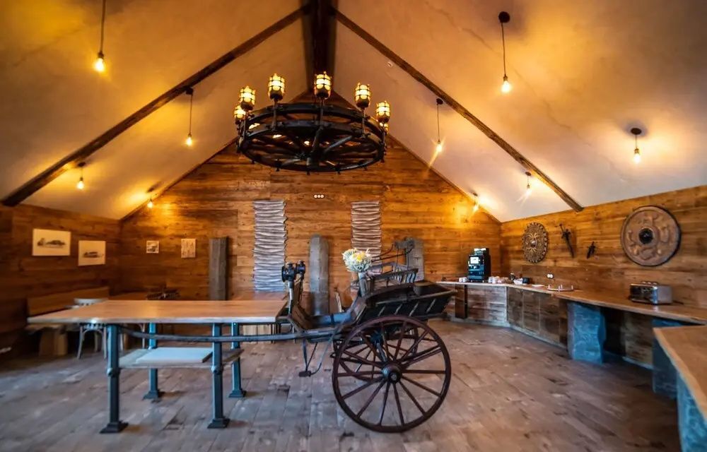 Rustikaler Innenraum aus Holz mit antikem Holzwagen und großem Radkronleuchter unter dem Giebel.