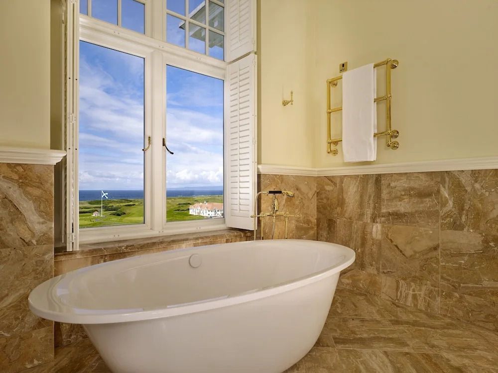 Freistehende Badewanne mit Blick auf das Meer durch ein Fenster, beige Fliesen