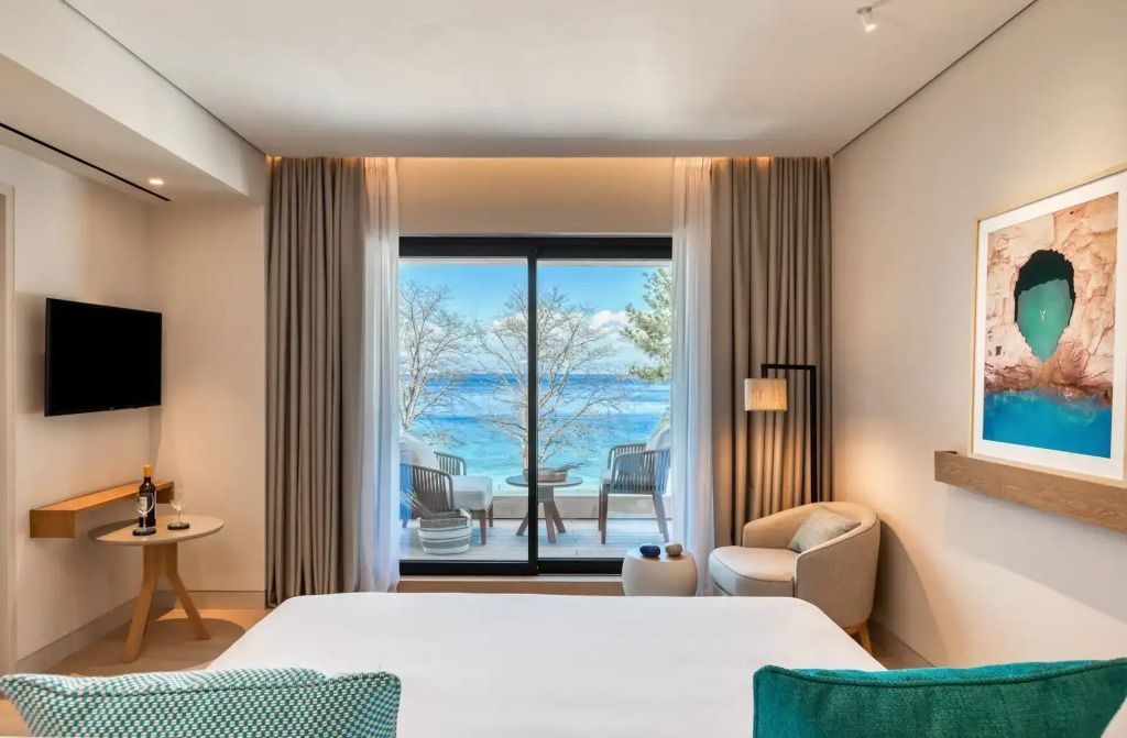Zimmer mit Bett und direktem Blick auf das Meer durch große Fenster.