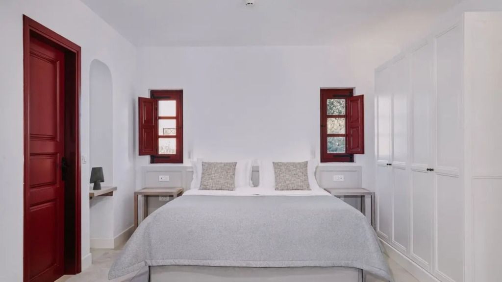 Gemütliches Schlafzimmer mit Doppelbett und roten Fensterläden im typischen Santorini-Stil.