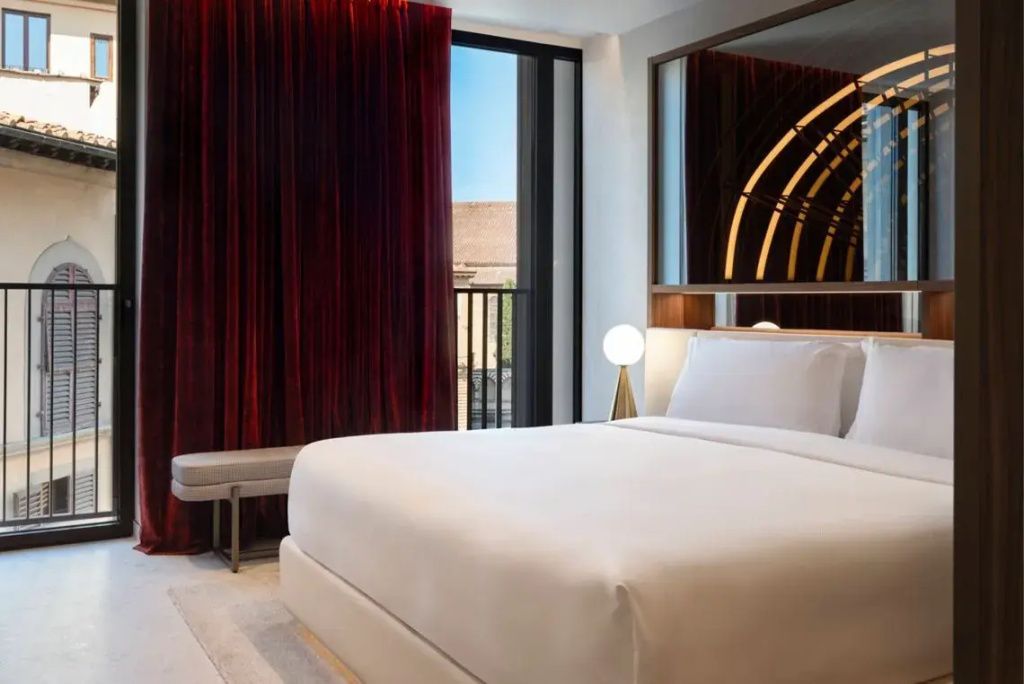 Modernes Schlafzimmer mit luxuriöser Bettwäsche, großer Spiegel, rote Vorhänge und Fenster mit Balkonblick.