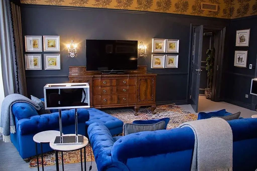 Wohnbereich mit blauen Sofas und antikem Holzschrank vor einem großen Fernseher