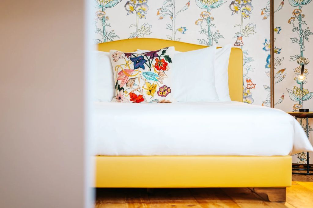 Bett mit floralem Kissen vor gemusterter Tapete und gelbem Rahmen