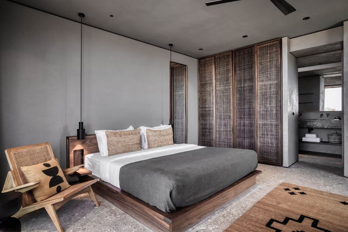 Doppelbett mit grauer Tagesdecke, Kissen und Schrank mit geflochtenen Fronten