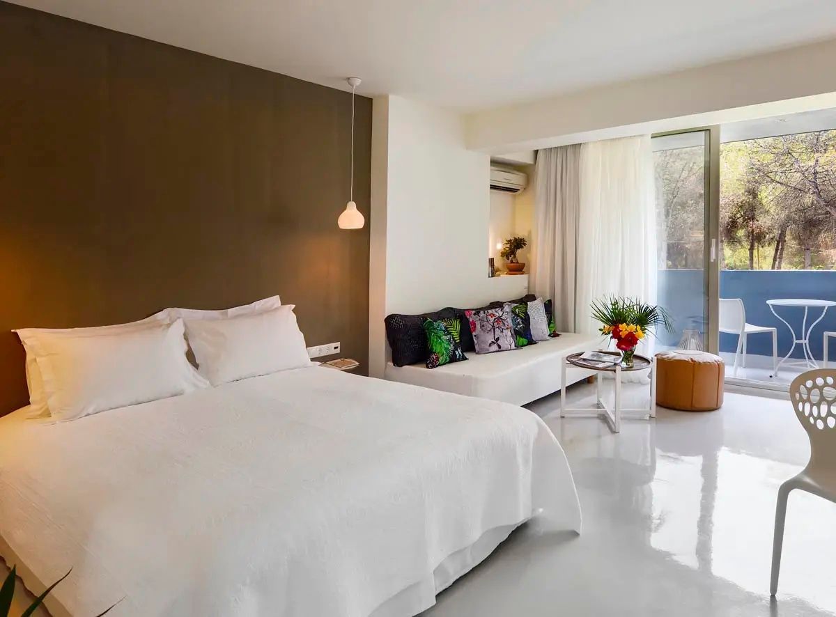 Doppelbett mit weißer Bettwäsche, brauner Wand und weißem Sofa mit bunten Kissen im Hotelzimmer.