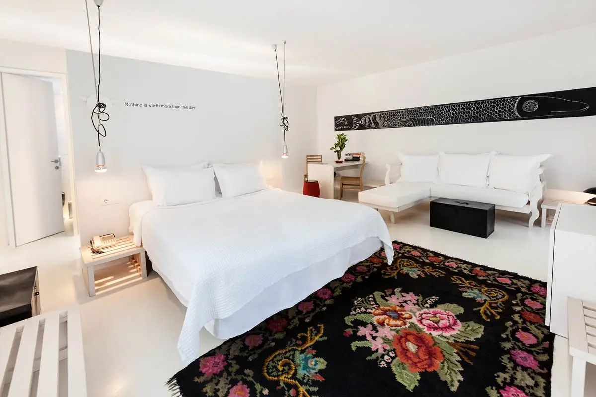 Weißes Hotelzimmer mit Doppelbett, hellem Sofa, Schreibtisch und buntem Teppich mit Blumenmuster.