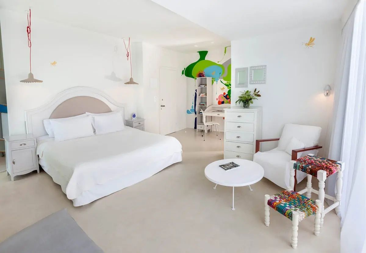 Weißes Schlafzimmer mit Sessel, kleinem runden Tisch, bunten Hockern und farbenfroher Wandzeichnung.