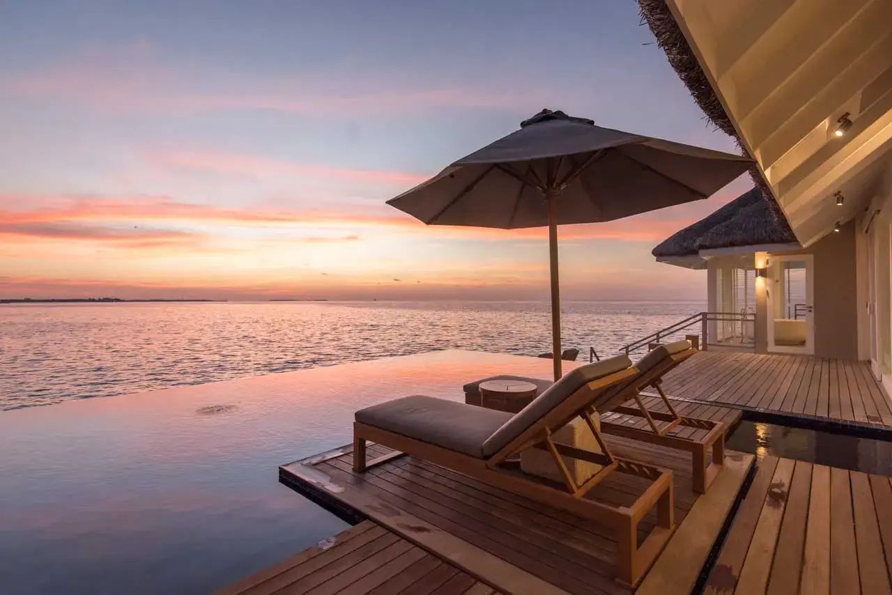Sonnendeck einer Wasservilla mit Sonnenliegen, Sonnenschirm und Infinity Pool im Abendlicht.