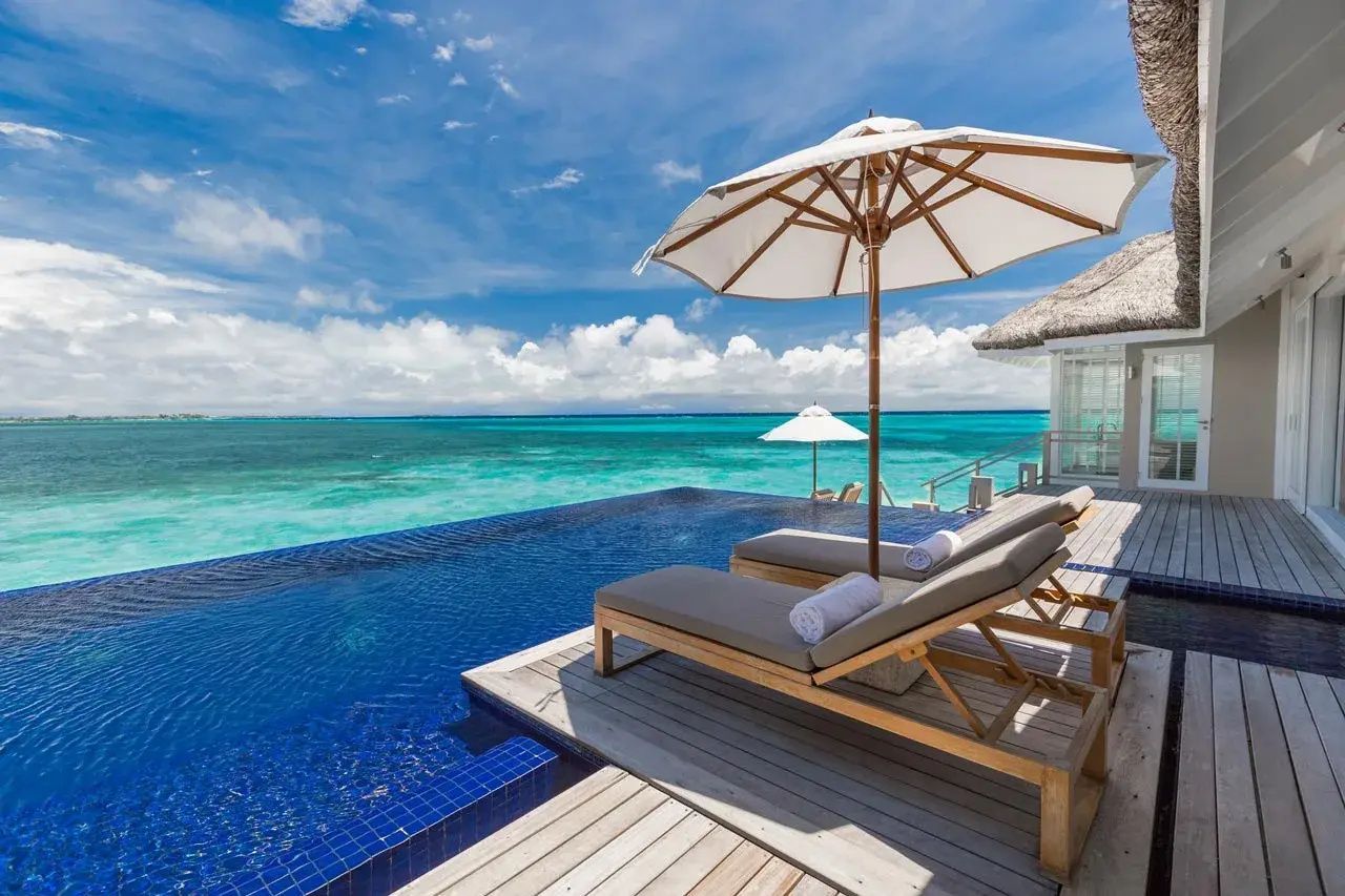 Infinity Pool auf Holzdeck mit zwei Liegen und Sonnenschirm mit Blick auf das Meer.