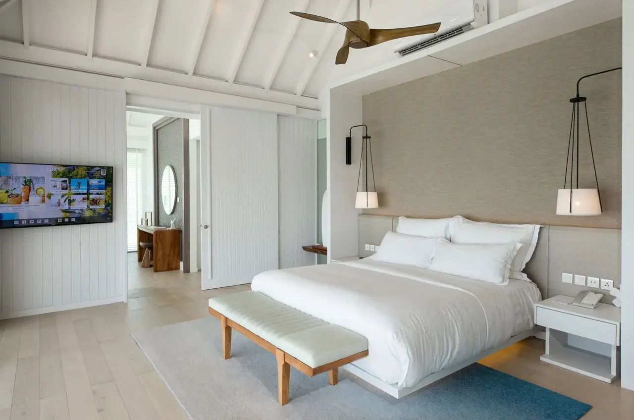 Heller Schlafbereich einer Villa mit Doppelbett, Deckenventilator und Schiebetür zum Bad.