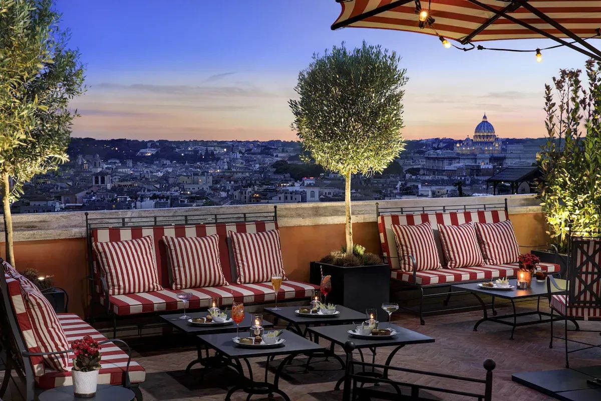 Elegante Dachterrasse des Rocco Forte Hotel de la Ville in Rom mit gestreiften Sitzpolstern, Cocktailtischen und Blick auf die Stadt sowie den Petersdom bei Sonnenuntergang.
