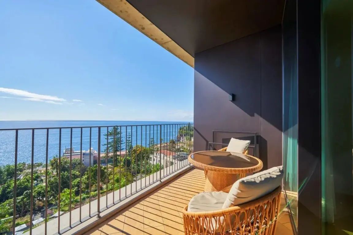 Überdachter Balkon mit Sitzgruppe aus Rattan und Blick auf das blaue Meer und die Küste.