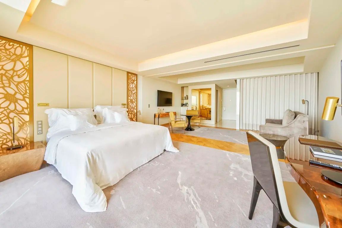 Hell gestaltetes Hotelzimmer mit großem Bett, Flachbildfernseher und Sitzbereich mit zwei Sesseln.