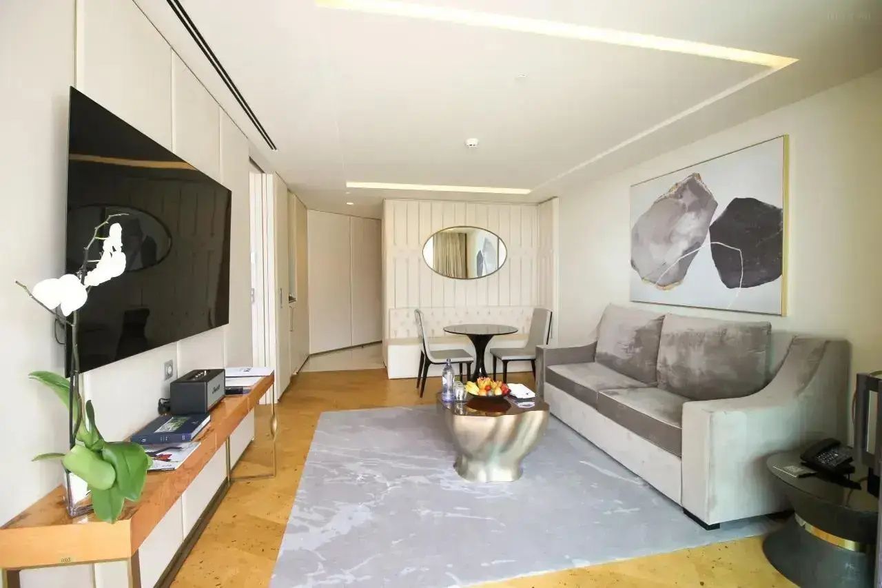 Wohnbereich der Suite mit grauem Sofa, Couchtisch, Essplatz und Fernseher an der Wand.