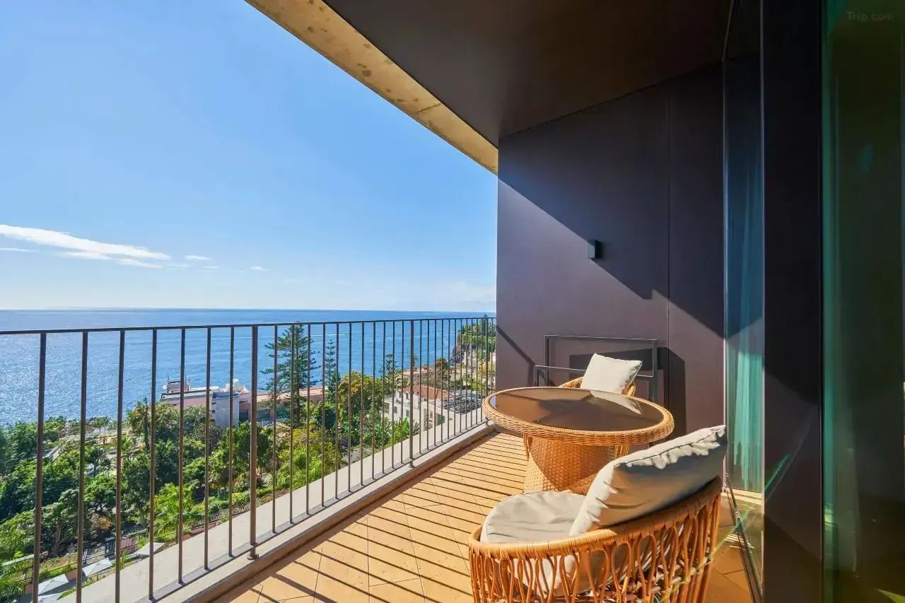 Überdachter Balkon mit Sesseln, Tisch und Blick auf das blaue Meer sowie die grüne Küste.