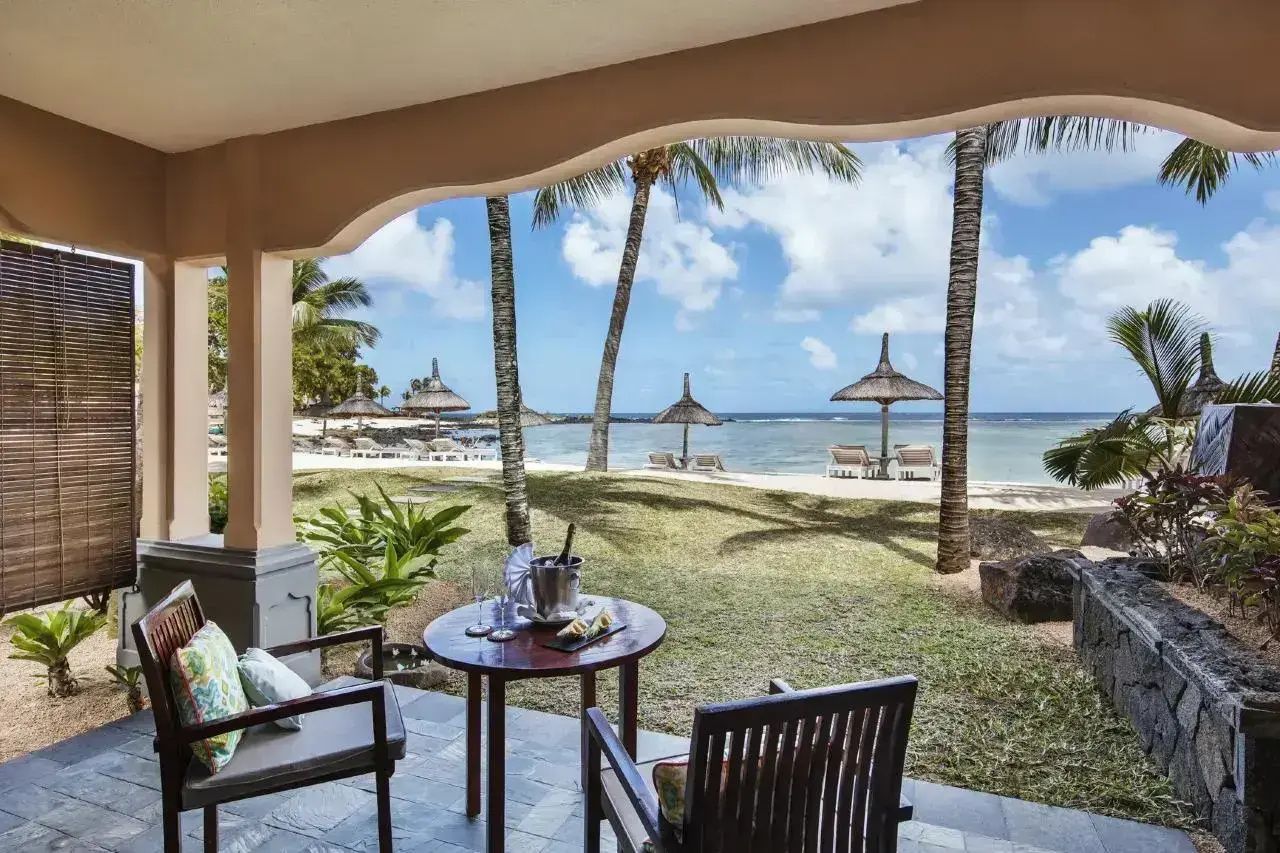 Blick von der überdachten Terrasse auf den Rasen, Palmen und Strand.