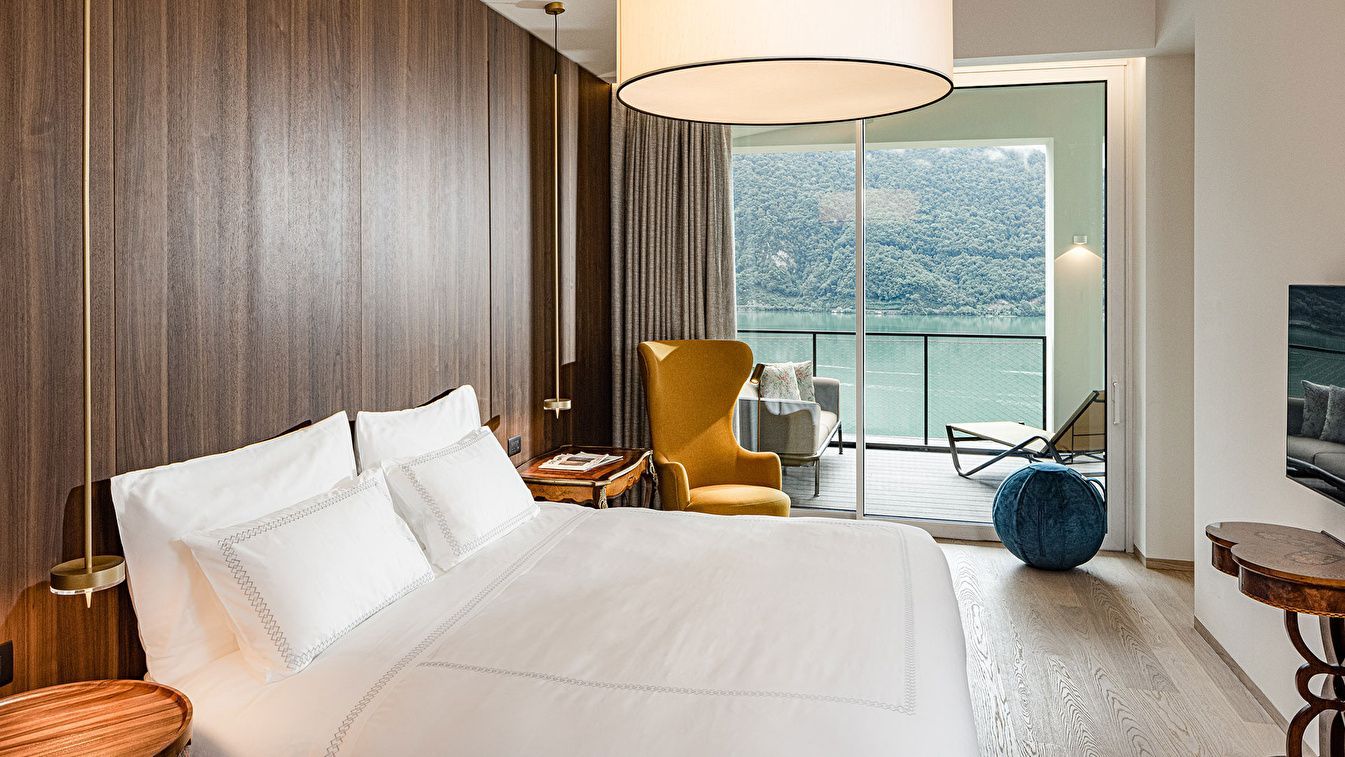 Hotelzimmer mit weißem Bett, gelbem Sessel und Glasfront mit Blick auf den See.