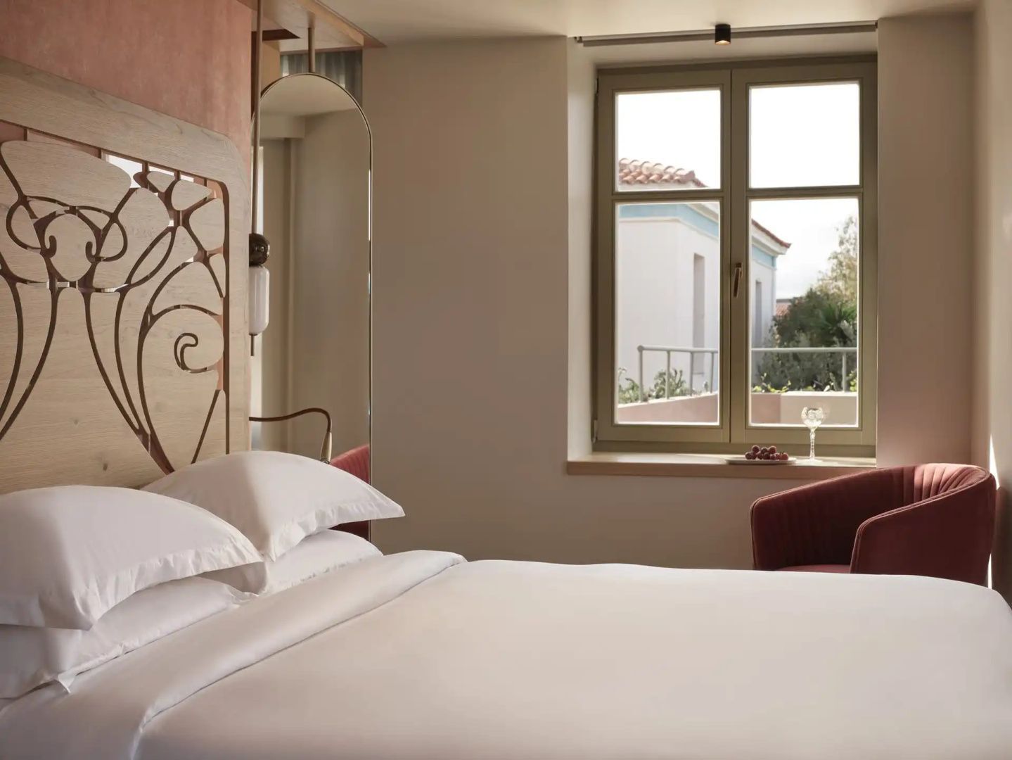 Doppelbett mit weißer Bettwäsche, verziertem Kopfteil und Blick durch das Fenster nach draußen.