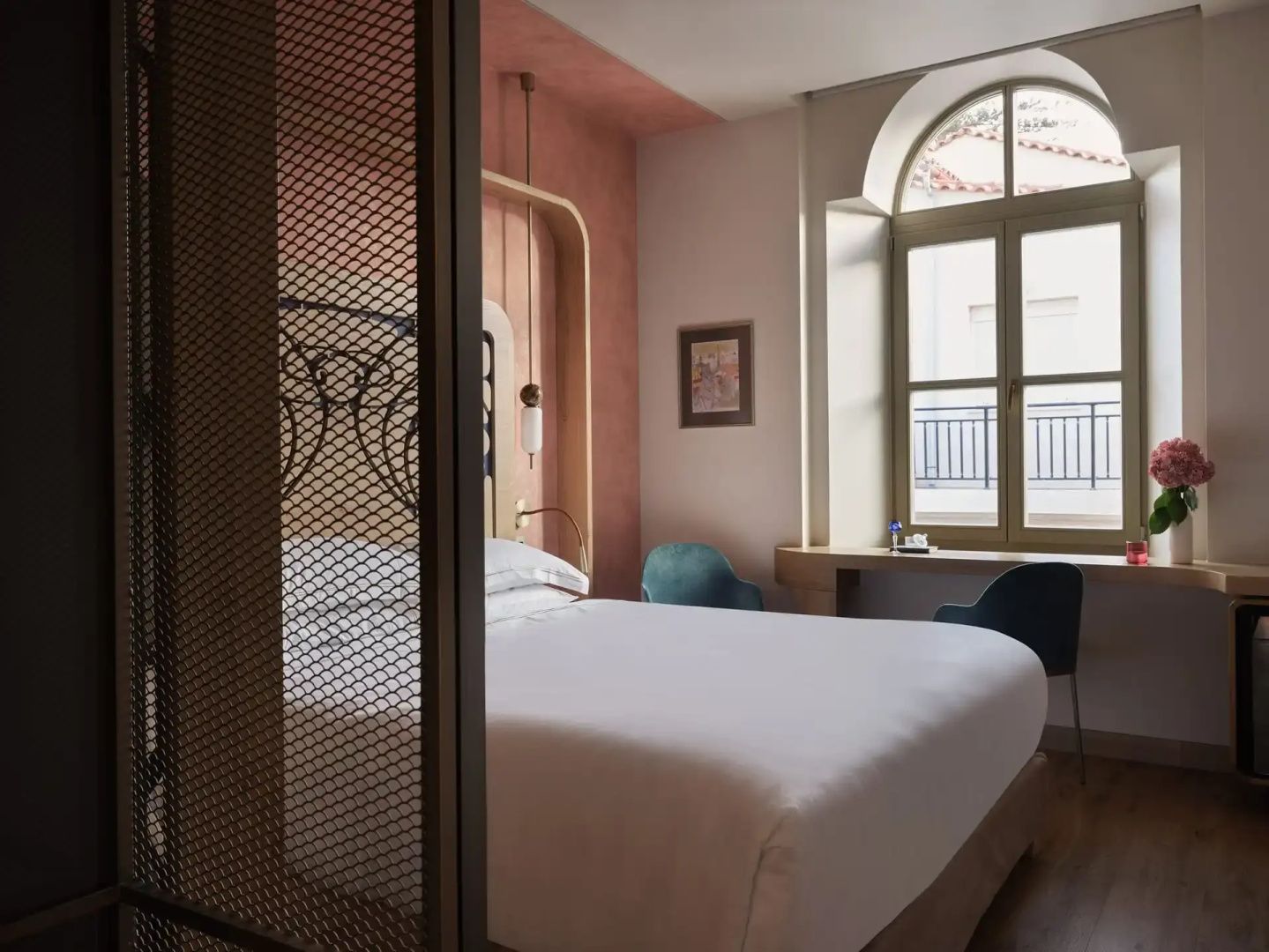 Ein eingerichtetes Hotelzimmer mit Doppelbett, rosa Wand, Raumteiler und einem Rundbogenfenster.