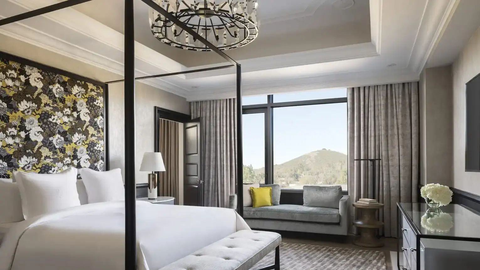 Schlafzimmer einer Suite mit weissem Himmelbett, Sitzecke am Fenster und Blick auf die Hügel.