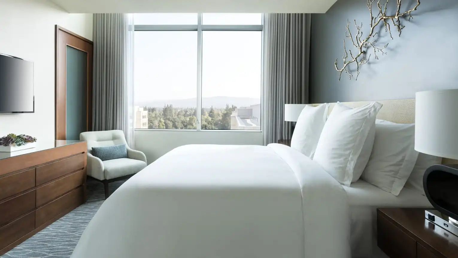 Helles Hotelzimmer mit weiß bezogenem Doppelbett, Sessel und Blick durch das Fenster.